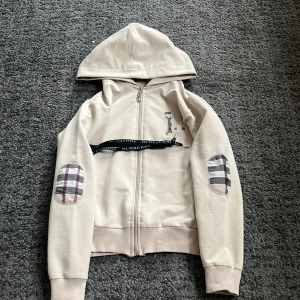 Beige huvtröja från Burberry - Säljer en snygg beige huvtröja från Burberry med dragkedja och klassiskt rutmönster på armbågarna. Tröjan har en broderad logotyp på bröstet och ribbade muddar. Perfekt för en stilren look!