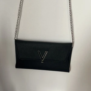 Svart kuvertväska från Valentino - Elegant svart kuvertväska från Valentino med en stilren design. Väskan har en kedjerem och ett stort V i metall på framsidan. Perfekt för att bära det nödvändigaste med stil.
