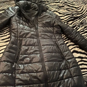 Svart dunjacka från Calvin Klein - Säljer en snygg svart dunjacka från Calvin Klein. Jackan har en quiltad design med dragkedja och huva. Perfekt för kyliga dagar och ger en stilren look. Tillverkad av återvunnet material för en hållbar stil. Det står att den är storlek XL men passar M perfekt!