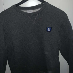 Grå sweatshirt från Les Deux - Säljer en stilren grå sweatshirt från Les Deux med ett blått emblem på bröstet. Tröjan har långa ärmar och en rund halsringning. Perfekt för en avslappnad stil. Den är i bra skick