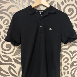 Svart pikétröja från Lacoste - Snygg svart pikétröja från Lacoste med klassisk krage och knappslå. Perfekt för en stilren look. 🐊Skriv för fler bilder, org pris 1400kr