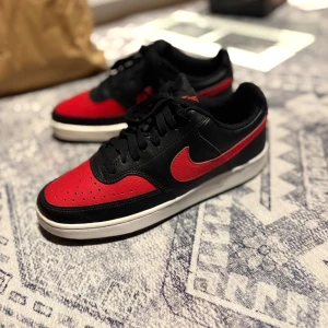 Nike Court Vision  - Snygga Nike Court Vision sneakers i svart och röd. Skorna har en klassisk design med svart ovandel och röda detaljer, inklusive den ikoniska Swoosh-loggan. Perfekta för en stilren look men även till att träna