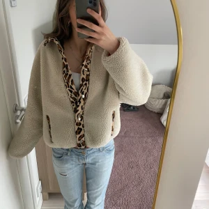 Beige teddyjacka med leoparddetaljer - Säljer en mysig beige teddyjacka med leopardmönstrade detaljer. Jackan har en loose passform och långa ärmar, perfekt för kyliga dagar. Den har en öppen design utan dragkedja eller knappar.