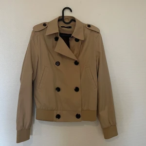 Beige trenchcoat från Saints de Mortals - Beige trenchcoat från Saints de Mortals med dubbelknäppning och axelklaffar. Jackan har långa ärmar och ribbade muddar vid ärmslut och nederkant.