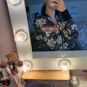 Blommig tröja från Adidas - Säljer en snygg blommig tröja från Adidas med dragkedja och svartvitt mönster. Jackan har långa ärmar och en sportig look med märkets logga på bröstet. Perfekt för en trendig stil! 💐