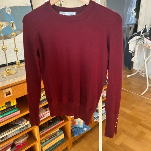 Vinröd tröja från Zara - Säljer en stilren vinröd tröja med knappar på ärmarna från Zara. Tröjan har långa ärmar och en rund halsringning. Perfekt för både vardag och mer uppklädda tillfällen. Materialet är mjukt och bekvämt.