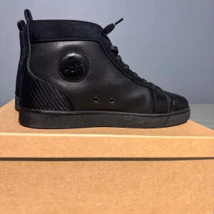 Svarta sneakers från Christian Louboutin - Snygga svarta sneakers från Christian Louboutin med perforerade detaljer och den ikoniska röda sulan. Skorna har snörning och en diskret logotyp på sidan. Perfekta för dig som vill ha en stilren look med en touch av lyx.