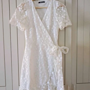Vit spetsklänning från Gina Tricot - Säljer en vacker vit spetsklänning från Gina Tricot. Klänningen har en omlott-design med knytband i midjan och korta ärmar. Perfekt för studenten eller sommaren. 🌸