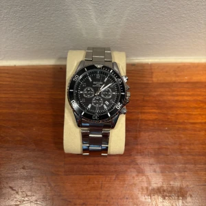 Silverfärgad klocka med svart urtavla - Snygg silverfärgad klocka med svart urtavla och kronografdetaljer. Klockan har en robust metallarmband och tydliga markeringar på bezeln. Perfekt för den som gillar en stilren och elegant look. 