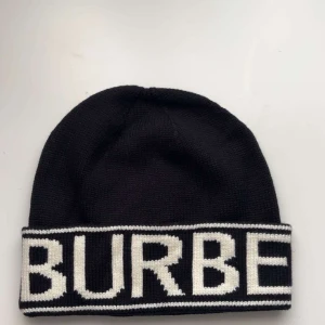 Burberry mössa - Snygg svart stickad mössa från Burberry med vit text runt kanten med prislapp på.