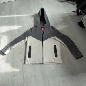 Grå och vit hoodie från Nike - Säljer en snygg grå och vit hoodie från Nike med dragkedja och huva. Jackan har två praktiska fickor med dragkedjor framtill och Nike-logga på bröstet. Perfekt för en sportig look!