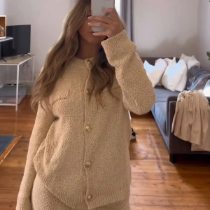 Guldig stickad kofta med knappar - Säljer en mysig beige guld stickad kofta med knappar framtill och två dekorativa fickor. Koftan har långa ärmar och ribbade muddar vid ärmslut och nederkant. Perfekt för kyliga dagar!