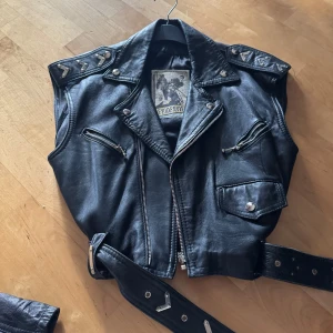 Svart skinnväst  - Cool svart skinnväst från Ed Rider med nitar på axlarna och flera dragkedjor. Västen har en klassisk bikerstil med bälte i midjan storlek 36