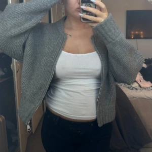 Grå ribbad kofta med dragkedja - Säljer en stilren grå kofta med dragkedja framtill. Den är ribbad och har långa ärmar, perfekt för kyligare dagar. Koftan har en enkel och modern design som passar till det mesta. Perfekt för lager-på-lager-stil.