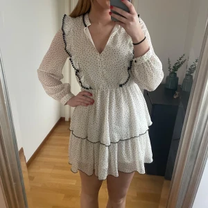 Vit prickig klänning med volangdetaljer - Vit klänning med svarta prickar och volangdetaljer. Den har en v-ringad design och långa ärmar. Perfekt till sommaren!
