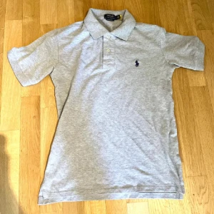 Grå pikétröja från Ralph Lauren - Säljer en stilren grå pikétröja från Ralph Lauren med klassisk krage och knappar. Tröjan har en broderad logga på bröstet och är kortärmad. Perfekt för en avslappnad men ändå snygg look.