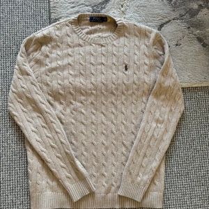 Beige kabelstickad tröja från Ralph Lauren - Säljer en stilren beige kabelstickad tröja från Ralph Lauren. Tröjan har en klassisk rund hals och det ikoniska logotypbroderiet på bröstet. Perfekt för en elegant och avslappnad look.