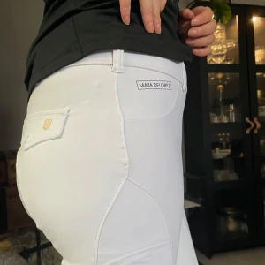 Vita ridbyxor från Maya Delorez - Snygga vita ridbyxor från Maya Delorez med diskret mönster och logotypdetaljer. Byxorna har en tight passform och är tillverkade i ett stretchigt material för optimal rörelsefrihet. Perfekta för ridning med stilren design och praktiska fickor.