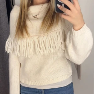 Vit stickad tröja med fransar - Säljer en mysig vit stickad tröja med fransdetaljer över bröstet. Tröjan har långa ärmar och en lös passform, både stilren och trendig, ett måste i garderoben!