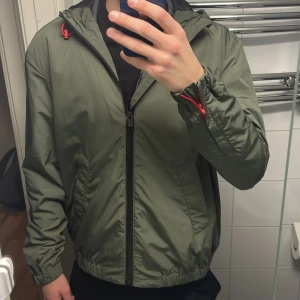 Grön vindjacka från Zadig & Voltaire - Snygg grön vindjacka från Zadig & Voltaire med dragkedja och huva. Jackan har röda detaljer vid dragkedjorna och är perfekt för blåsiga dagar. Den har en normal passform och är tillverkad i ett lätt syntetmaterial. Tags medföljer då den är helt ny! Lägg prisförslag så jobbar vi där ifrån.