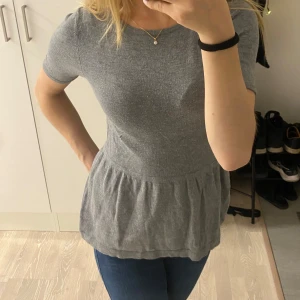 Grå kortärmad topp - Säljer en stilren grå topp med korta ärmar och en lös passform. Toppen har en volangdetalj vid nederkanten som ger en fin touch. Perfekt för en avslappnad look.