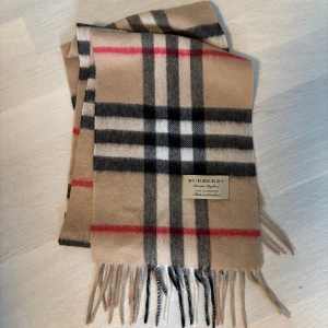 Beige rutig halsduk från Burberry - Säljer en klassisk beige rutig halsduk från Burberry med fransar i ändarna. Den har inslag av svart, vit och röd i mönstret. Perfekt för att hålla sig varm och stilren under kyliga dagar.
