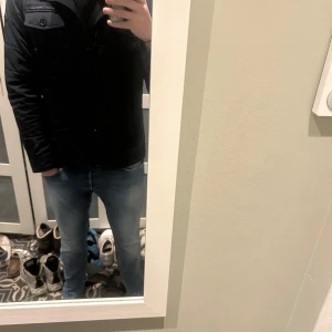 J Lindberg jacka - Säljer en stilren svart parkajacka med dragkedja och knappar. Jackan har långa ärmar och en klassisk design som passar perfekt för kyligare dagar. Perfekt för dig som vill ha en tidlös look.