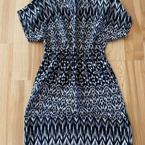 Svartvit mönstrad klänning XS - Snygg kortklänning i storlek XS med svartvitt grafiskt mönster. Klänningen har korta ärmar och resår i midjan för en skön passform. Tillverkad i mjuk polyester som känns lätt mot huden. Perfekt för dig som gillar statement-plagg med coola mönster.