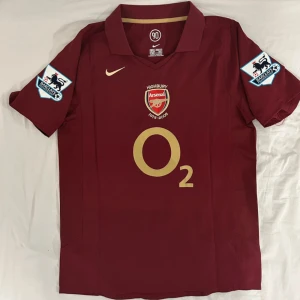 Arsenal Highbury Henry 14 Nike tröja - Snygg Arsenal Highbury 1913-2006 matchtröja i vinrött med guldiga detaljer och O2 sponsortryck. Kortärmad modell med klassisk krage, Premier League-patchar på ärmarna och Henry 14 på ryggen. Perfekt för dig som älskar fotbollshistoria och ikoniska spelare.
