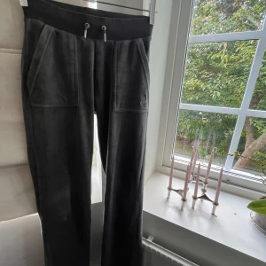 Mörkgrå mjukisbyxor Juicy Couture - Mörkgrå mjukisbyxor från Juicy Couture i mjuk velour med bred resårmidja och snörning. Byxorna har stora fickor fram och bak med silverfärgade knappar och diskret JC-logo. Perfekta för chill dagar och snygg streetstil. Se skada sista bilden