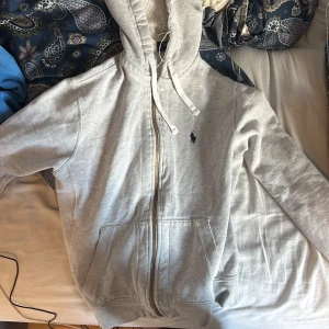 Grå hoodie från Polo Ralph Lauren - Snygg ljusgrå hoodie från Polo Ralph Lauren med dragkedja, huva med snörning och klassisk blå logga på bröstet. Tillverkad i mjuk bomull och har två stora fickor framtill. Perfekt för chill dagar eller när du vill ha en clean look.