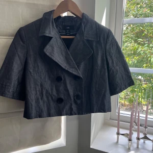 Kort grå kavaj från H&M - Kort kavaj från H&M i mörkgrått linneliknande material. Dubbelknäppt framtill med stora svarta knappar och bred krage. Modellen är boxig och har korta ärmar, perfekt för en trendig och avslappnad look.