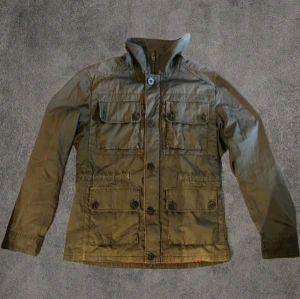 J.Lindeberg feild jacka - Den är mer grön i verkligheten det är konstigt ljus.Snygg olivgrön jacka från J.Lindeberg i storlek M. Jackan har fyra stora fickor framtill med knappar, hög krage och både dragkedja och knäppning. Perfekt för dig som gillar stilren och funktionell design. Perfekt inför hösten 👌Lite sliten längst ner vid midjan, men det är mer på insidan så det syns inte vid användning😊