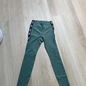 Gröna leggings från Stronger - Snygga gröna tränings byxor från Stronger med svarta sidor där det står 'STRONGER' i vitt. De har en tight passform och är gjorda i ett stretchigt material som passar perfekt till träning. Diskret ribbad struktur i tyget för extra stilpoäng.