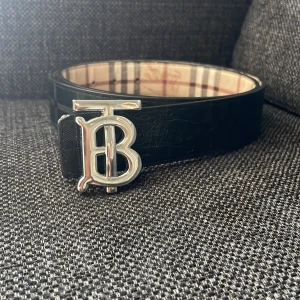 Burberry bälte - Snyggt Burberry-bälte i svart skinn med klassiskt rutigt mönster på båda sidor. Ena sidan är svart med präglat rutmönster och logga, andra sidan har beige, röd och grå rutor. Stor silverfärgad TB-spänne framtill. Perfekt accessoar för att lyfta din outfit.