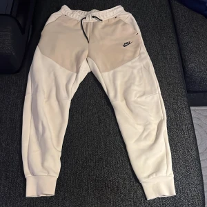 Nike mjukisbyxor i beige och vitt - Snygga mjukisbyxor från Nike i beige och vitt med svart snörning i midjan och logga på låret. Byxorna har elastisk midja, muddar vid bensluten och en dragkedjeficka bak. Perfekta för chill eller träning.