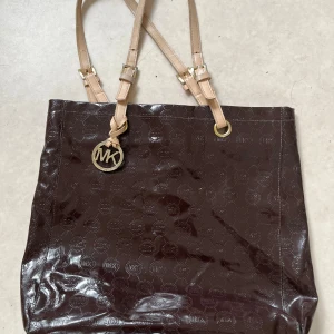 Brun Michael Kors axelväska i skinn - Snygg brun axelväska från Michael Kors med glansig finish och präglat MK-mönster över hela väskan. Väskan har ljusbeigea axelremmar med guldfärgade detaljer och ett MK-hänge framtill. Perfekt storlek för att få plats med allt du behöver.