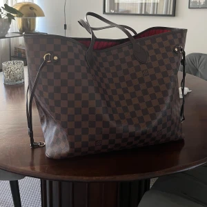 Damier Neverfullinspirerad väska - Säljer en stor och rymlig väska inspirerad av Neverfull med det klassiska brunrutiga Damier-mönstret. Väskan har långa handtag, guldfärgade detaljer och rödrandigt foder. Mini-clutch ingår. Fint skick!