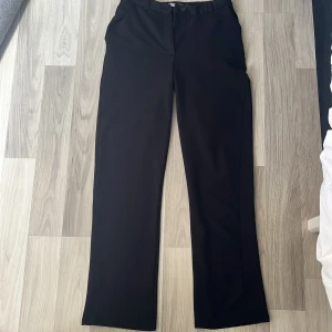 Svarta kostymbyxor i XS - Klassiska svarta kostymbyxor med raka ben och hällor för bälte. Byxorna har en enkel knappstängning och är perfekta för en clean och stilren look. Passar dig som gillar minimalistisk stil och vill ha ett par byxor som funkar till många outfits.