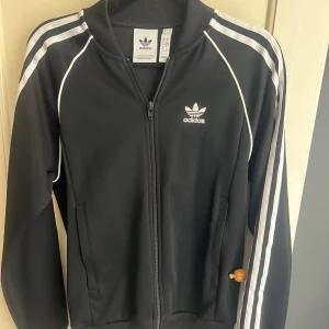 Svart Adidas bomberjacka med vita ränder - Klassisk svart Adidas med vita ränder längs ärmarna och vit. Ikonisk Adidas-logga på bröstet. Perfekt för en sportig och stilren look. Storlek xs.