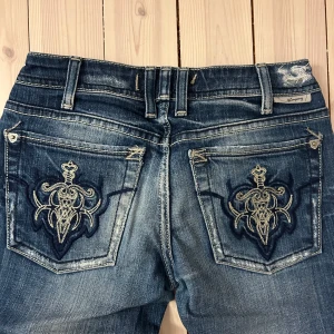 Lågmidjade bootcut jeans  - Lågmidjade bootcut jeans från Jcompany med broderier på bakgickorna, och slitna detaljer. Färgen är mellanblå med ljusare tvättade partier. De är i storlek 24, men de är dessutom avkortade. Säljer då de ej längre passar! 