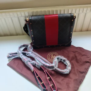 Svart och röd axelväska från Malini - Cool axelväska i svart skinn med röd bred rand och diskret rutmönster. Väskan har guldfärgade nitar längs kanterna och matchande detaljer på remmen. Insidan är beige och har flera fack, perfekt för att hålla ordning på grejerna. Snygg design med lyxig vibe.