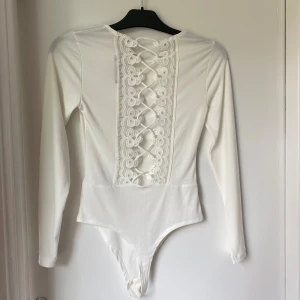 Vit långärmad body med spets från Bik Bok - Supersnygg vit body från Bik Bok med långa ärmar och spetsdetaljer längs ryggen. Baksidan har snörning och vackert mönstrad spets som ger en extra touch. Mjuk och stretchig, perfekt att styla med jeans eller kjol.