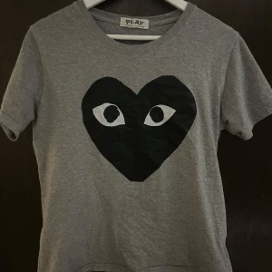 Grå t-shirt Comme des Garçons Play - Grå t-shirt från Comme des Garçons Play med klassiskt svart hjärta med ögon tryckt på bröstet. Rund hals och korta ärmar, tillverkad i mjuk bomull. Perfekt för dig som gillar streetwear och vill sticka ut med en ikonisk design.