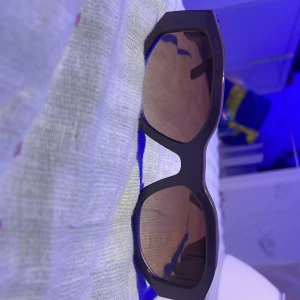 Bruna solglasögon med guldiga detaljer - Snygga solglasögon med svart, bred båge och bruna glas. Sidorna har unika guldiga metallringar som ger en cool och lyxig vibe. Perfekt accessoar för att sticka ut och ge din outfit en extra edge.