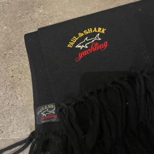 Svart halsduk från Paul & Shark - Svart halsduk från Paul & Shark Yachting med broderad logga i gult, vitt och rött. Klassisk design med långa fransar i ändarna. Tillverkad i Italien och perfekt för kalla dagar när du vill ha en stilren accessoar.
