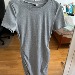 Grå ribbad kortklänning från H&M Divided - Snygg grå ribbad kortklänning från H&M Divided i storlek M. Klänningen har korta ärmar, rund hals och cool snörningsdetalj längs ena sidan som ger en schysst draperad effekt. Perfekt för dig som gillar enkel men trendig stil.