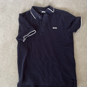Mörkblå BOSS pikétröja XS - Snygg mörkblå pikétröja från BOSS i storlek XS. Klassisk krage med vit kant och korta ärmar med vit piping. Tröjan har knappar vid halsen och BOSS-logga broderad på bröstet. Perfekt för en clean och stilren look.