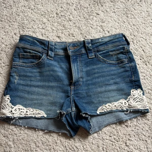 Blå jeansshorts med spets från H&M - Snygga blå jeansshorts från H&M med vit spetsdetalj på båda benen. Säljs på grund av att jag inte använder dom! Jeansshortsen är väldigt korta i Modellen. Fläckarna på baksidan är en del i designen då jag fick dom så när jag köpte dom.