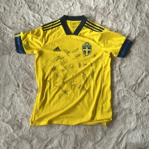 Svenska landslagets signerade matchtröja - Gul fotbollströja från Adidas med blå detaljer och Sveriges landslagsemblem på bröstet. Tröjan har korta ärmar, klassiska Adidas-ränder på axlarna och är tillverkad i lätt, ventilerande polyester. Flera autografer från svenska herrlandslaget på framsidan gör den extra unik för samlare. 
